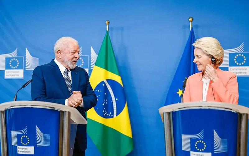 Dois líderes em pé atrás de púlpitos com bandeiras do Brasil e da UE ao fundo. Ambos sorriem em um cenário formal com azuis predominantes.