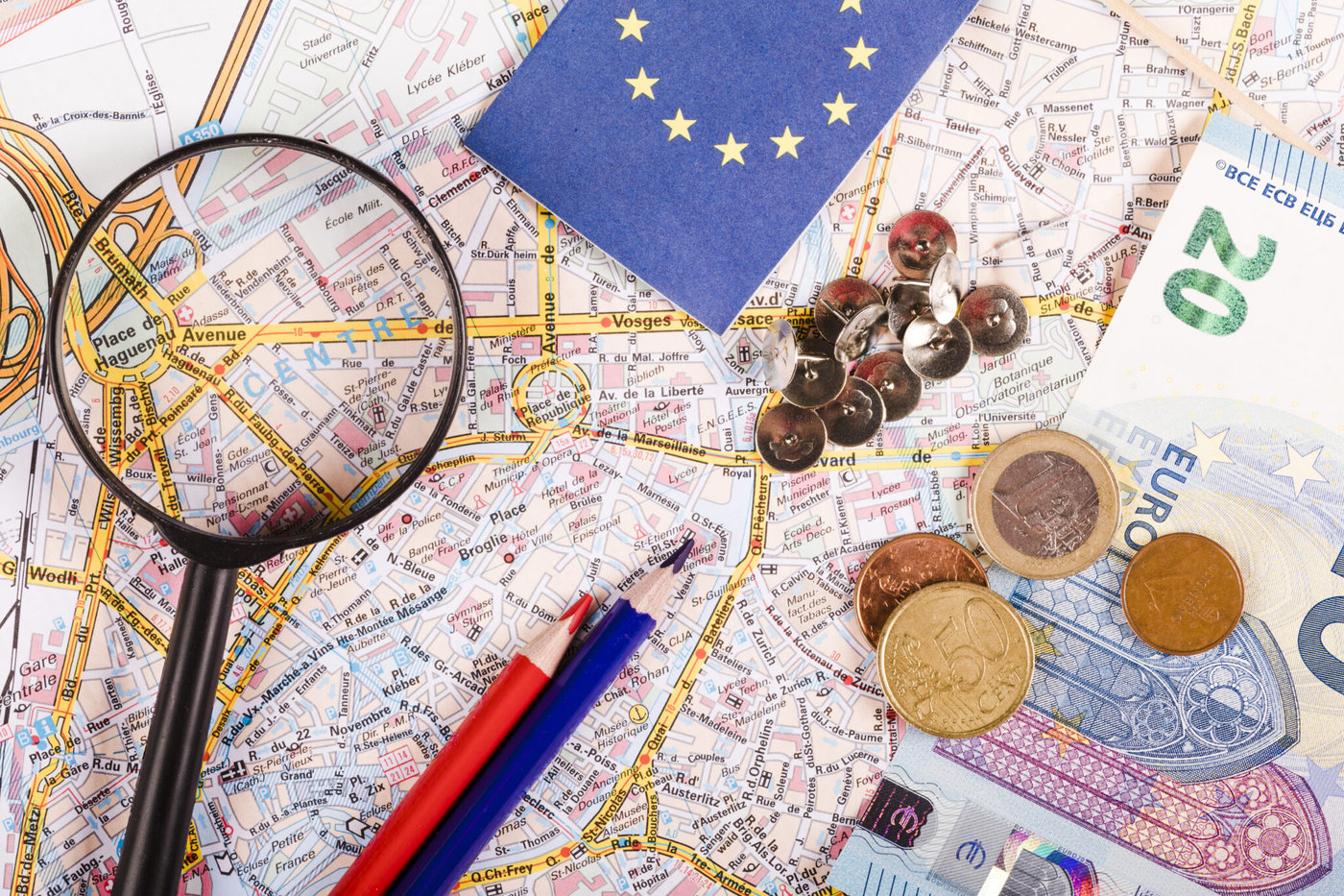 Mapa detalhado com lupa, tachas, lápis tricolor, moedas e cédula de 20 euros. Canto de bandeira da UE visível. Atmosfera de viagem.