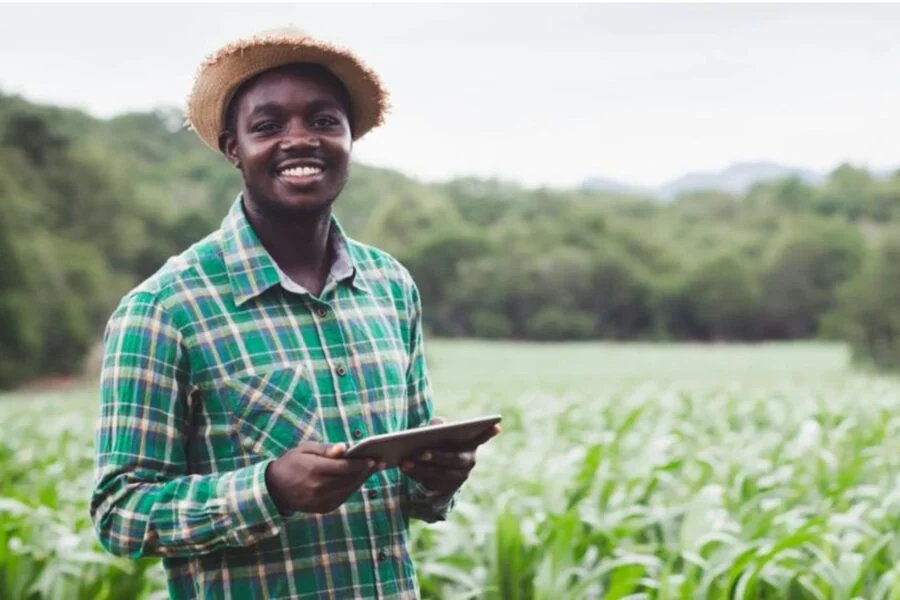 Homem sorridente de chapéu de palha e camisa xadrez verde, segurando um tablet em lavoura ao ar livre.