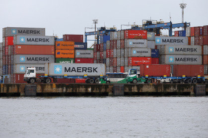 Porto com contêineres empilhados da Maersk, caminhões no cais e água calma em primeiro plano.