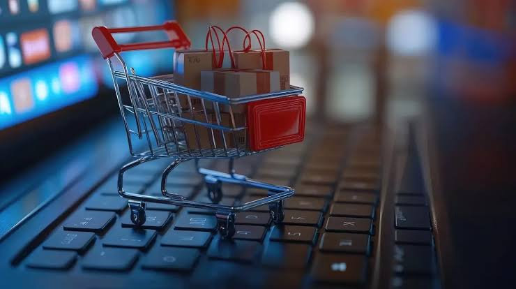 Mini carrinho de compras com pacotes sobre teclado de laptop, em cena de e-commerce com luzes desfocadas.