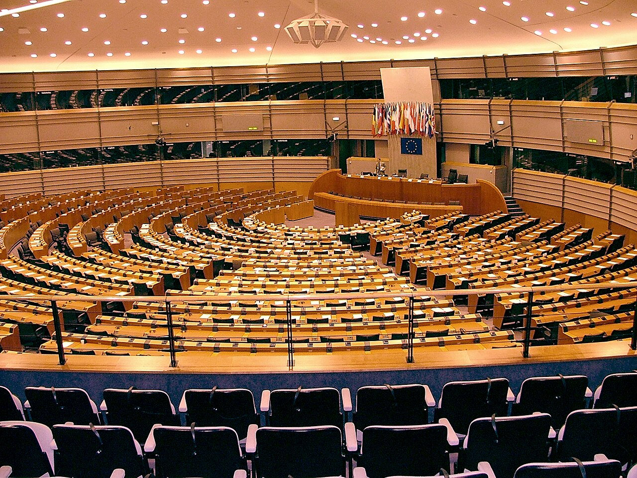Hemiciclo vazio do Parlamento Europeu com cadeiras circulares de madeira. Bandeiras dos países da UE e emblema europeu ao fundo.