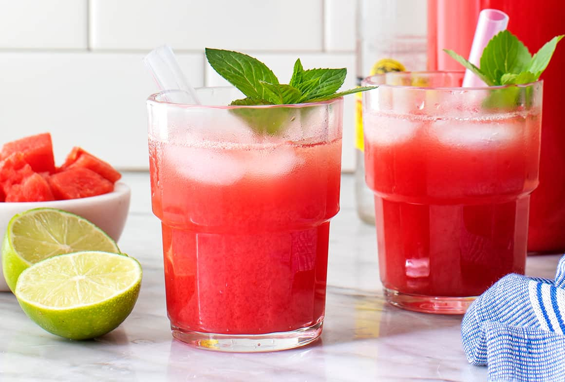 Duas bebidas vermelhas com gelo e hortelã sobre bancada, com limões e cubos de melancia ao lado.