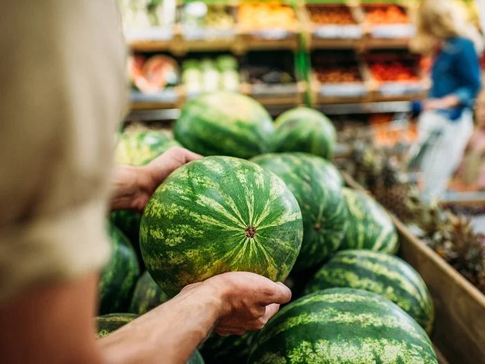 Pessoa segurando uma melancia em uma banca de frutas no supermercado, com outras melancias e prateleiras ao fundo.