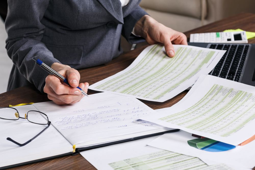 Pessoa de terno segura documentos com gráficos verdes em uma mesa, ao lado de notebook, óculos e calculadora. Ambiente de escritório.