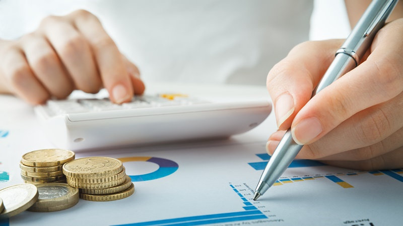 Mãos calculando e escrevendo em gráficos sobre mesa, com pilha de moedas ao lado. Fundo neutro, foco em finanças e planejamento.