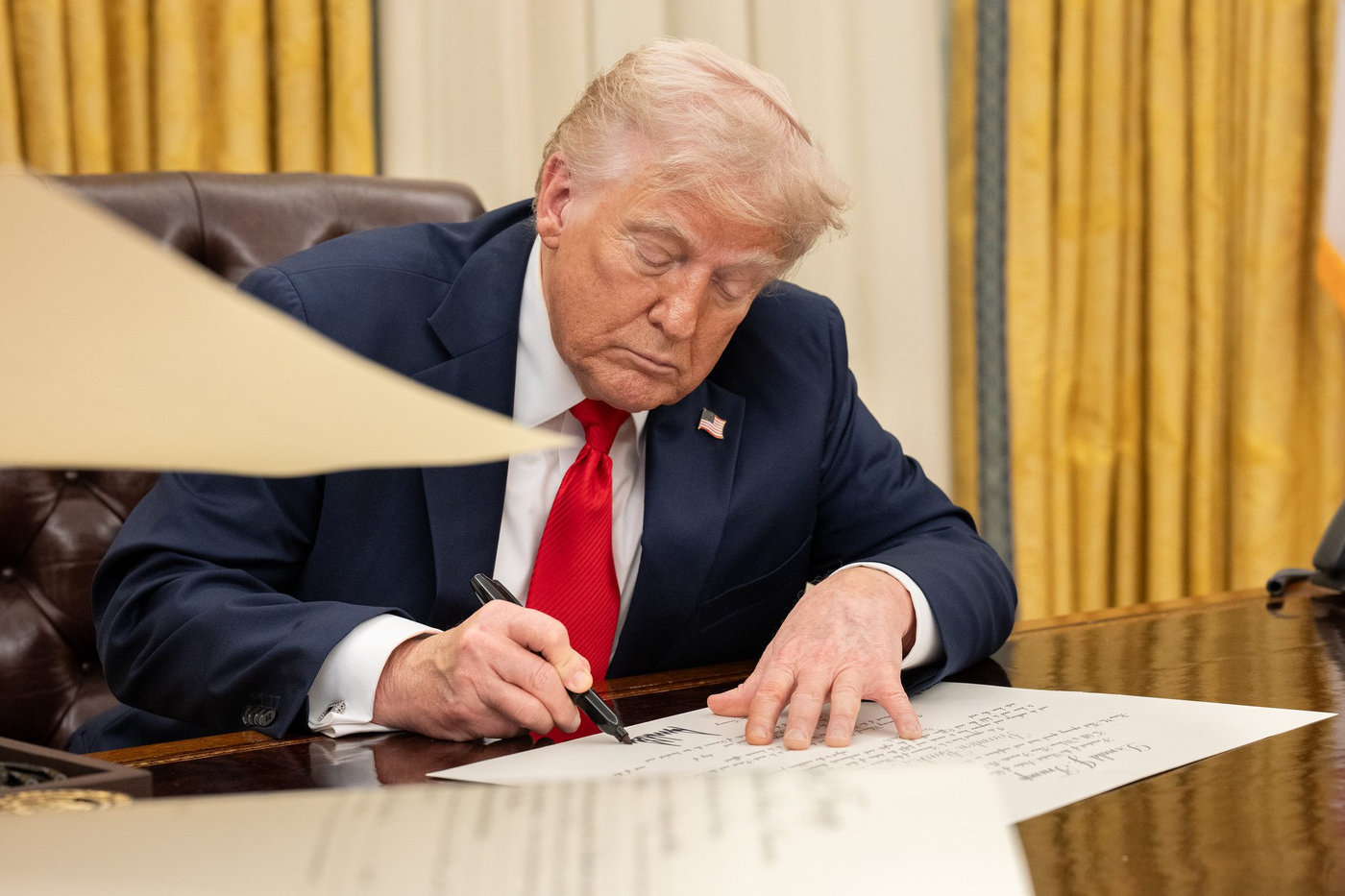 Presidente Donald Trump assinando documento sobre as tarifas em mesa de madeira. Cortinas douradas no fundo. Expressão concentrada. Papel à frente.